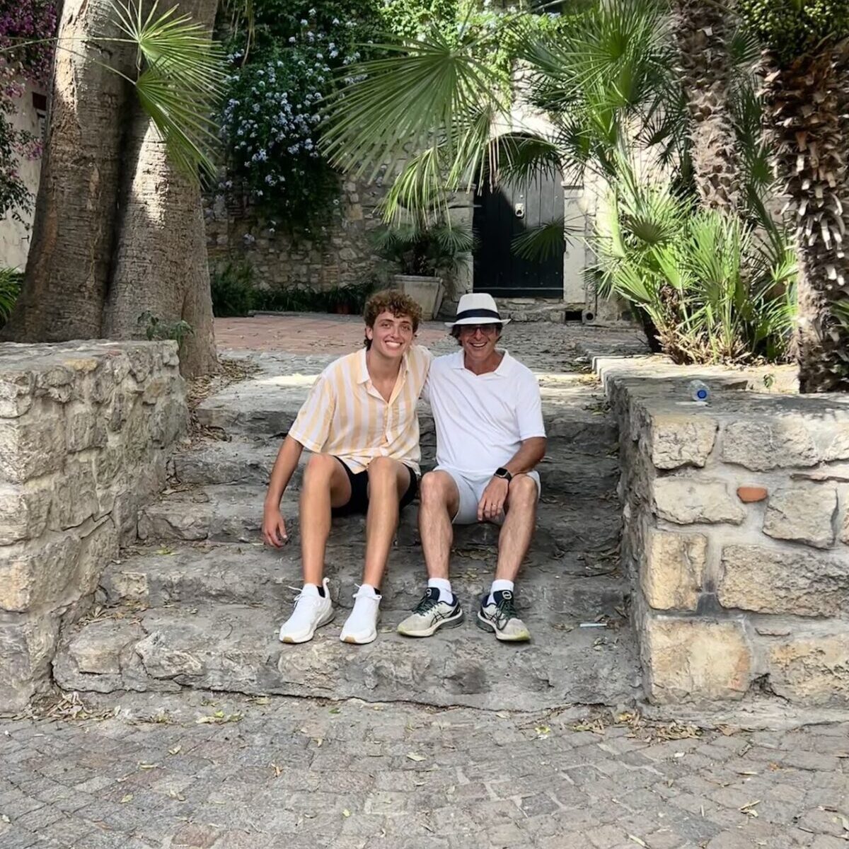 Two men sit on the steps with palm trees in the background.